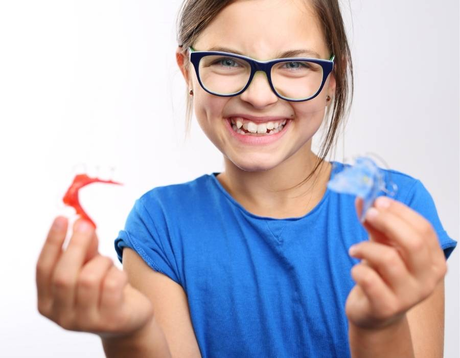 Smiling child holding two orthodontic retainers while wearing glasses.