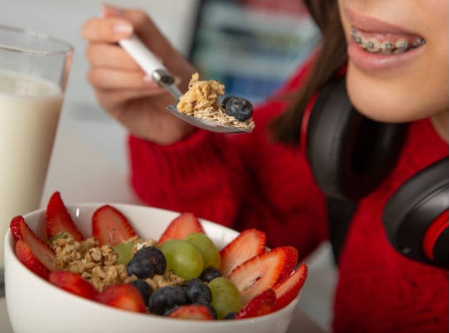 A person wearing metal dental braces and red headphones eating a healthy breakfast bowl of granola, strawberries, blueberries, and grapes.