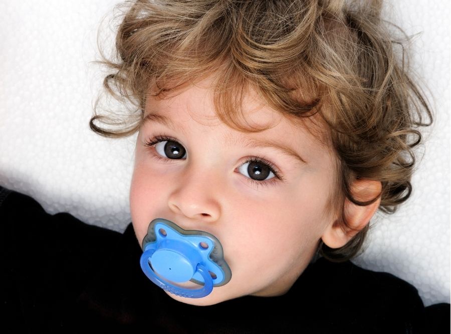 Close-up portrait of a curly-haired toddler laying down with a blue pacifier in their mouth